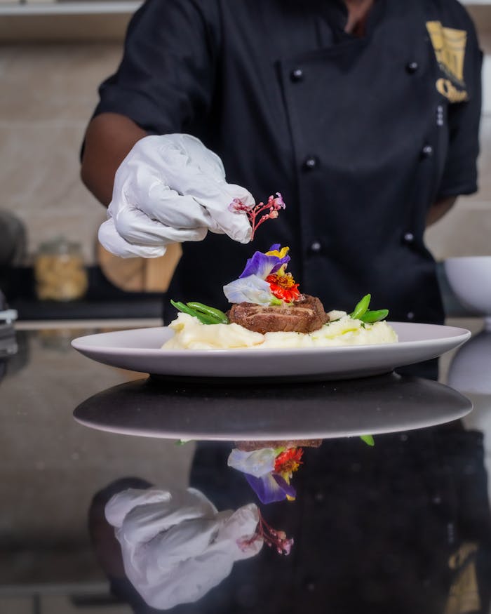 Chef carefully garnishing a gourmet beef steak with edible flowers and greens on a plate of mashed potatoes.